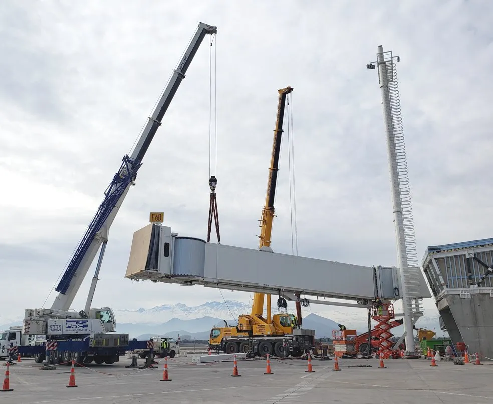 Two cranes lifting a boarding bridge tunnel during installation, with the Andes behind
